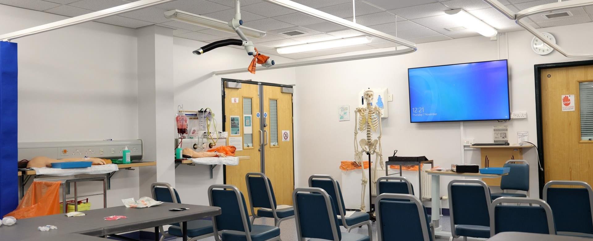 “A clinical teaching room with rows of chairs facing a screen on the wall. The room includes a skeleton model, medical equipment, and mannequins on side tables.”