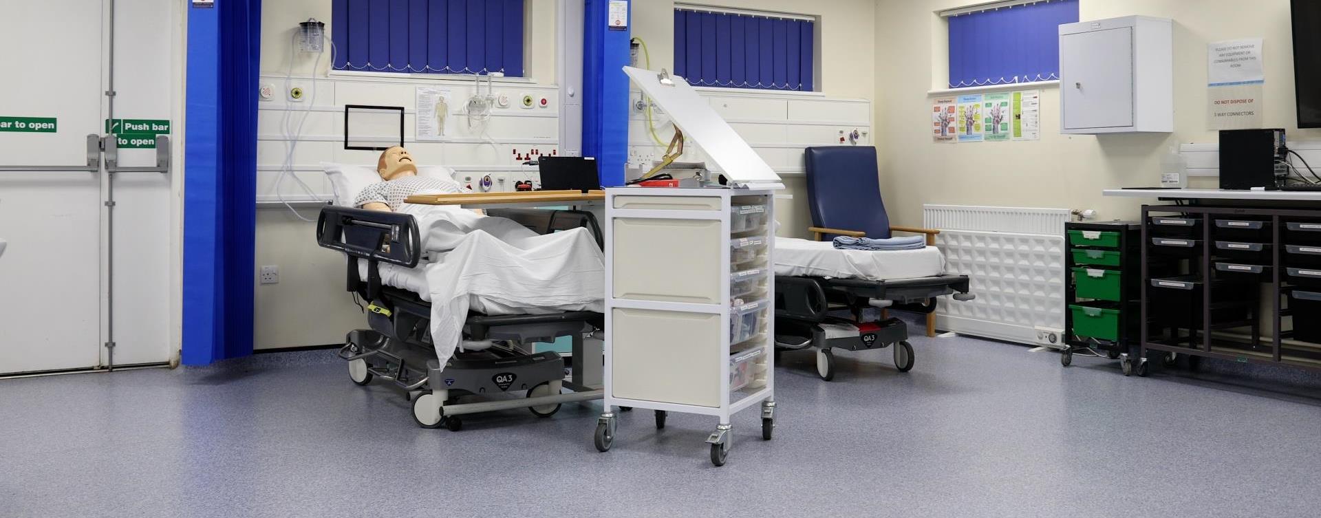 “A clinical simulation room featuring two hospital beds with mannequins, medical supplies on rolling carts, and mounted medical equipment along the walls.”