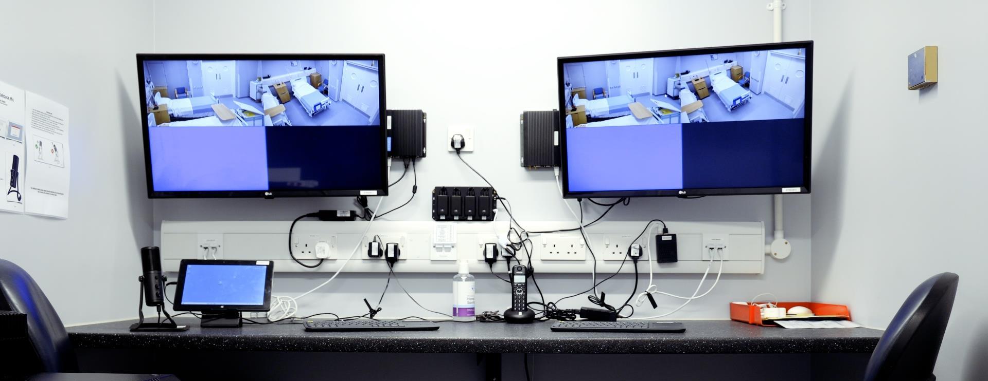 “Audio-visual control room with two large monitors displaying live feeds from a clinical training session. Equipment and wiring are arranged on the desk.”