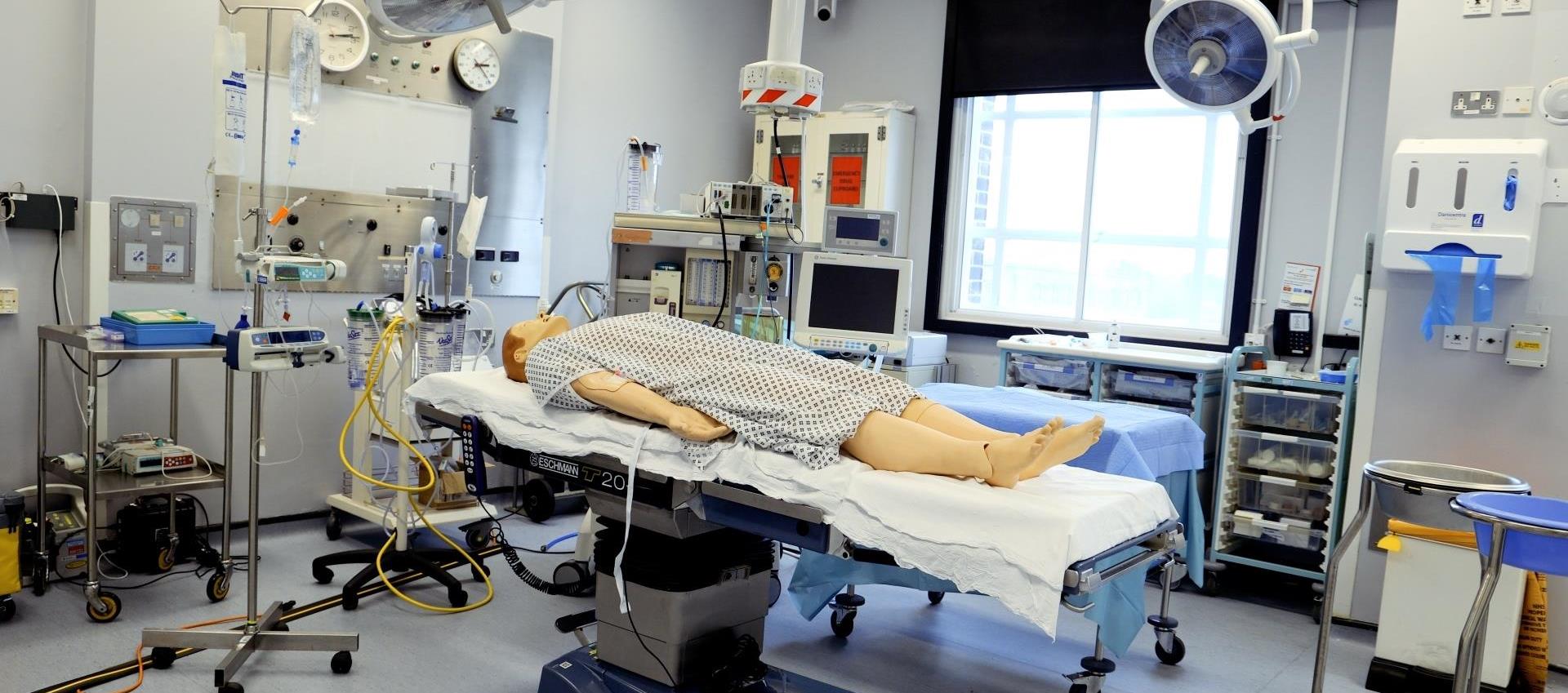 “A medical operating room simulation with a mannequin lying on a surgical table, surrounded by surgical lights, monitors, and medical equipment for practice.”