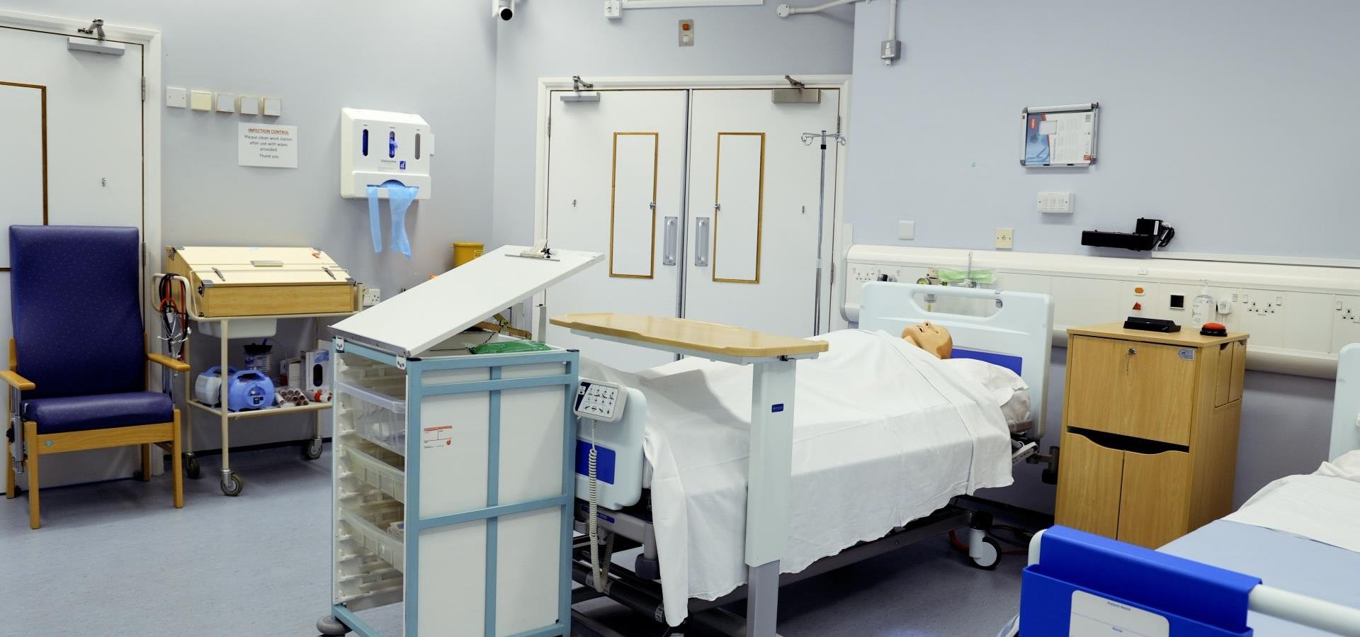 “Simulated hospital ward with a bed containing a mannequin, a rolling cart with medical tools, and infection control signs on the wall.”