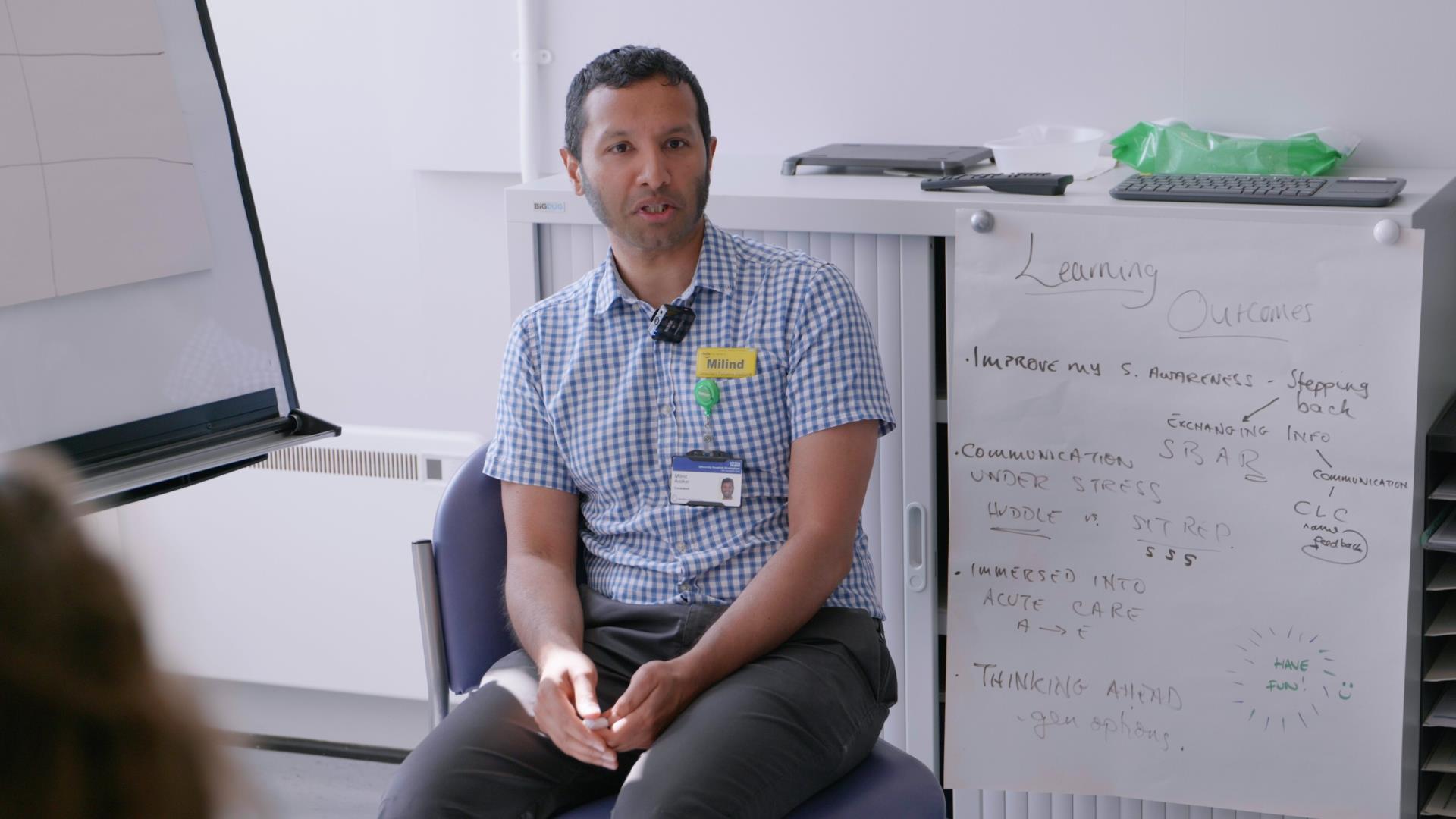 An instructor, seated in front of a whiteboard with ‘Learning Outcomes’ listed, leads a faculty development discussion. They wear a checked shirt with a visible name badge, engaging in conversation with the group