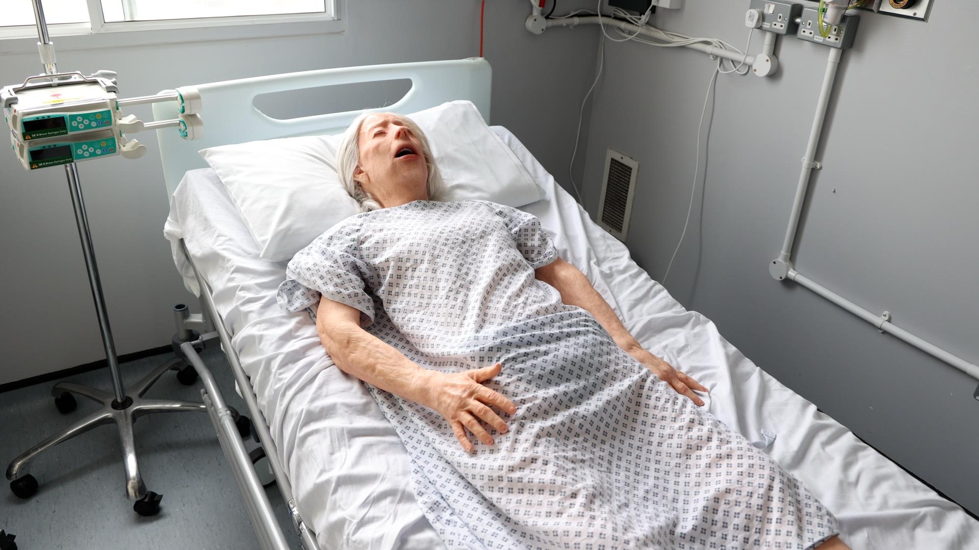 “A full-body view of a lifelike elderly mannequin lying on a hospital bed, wearing a patterned patient gown. Medical equipment, such as an IV pole, is present in the room.”