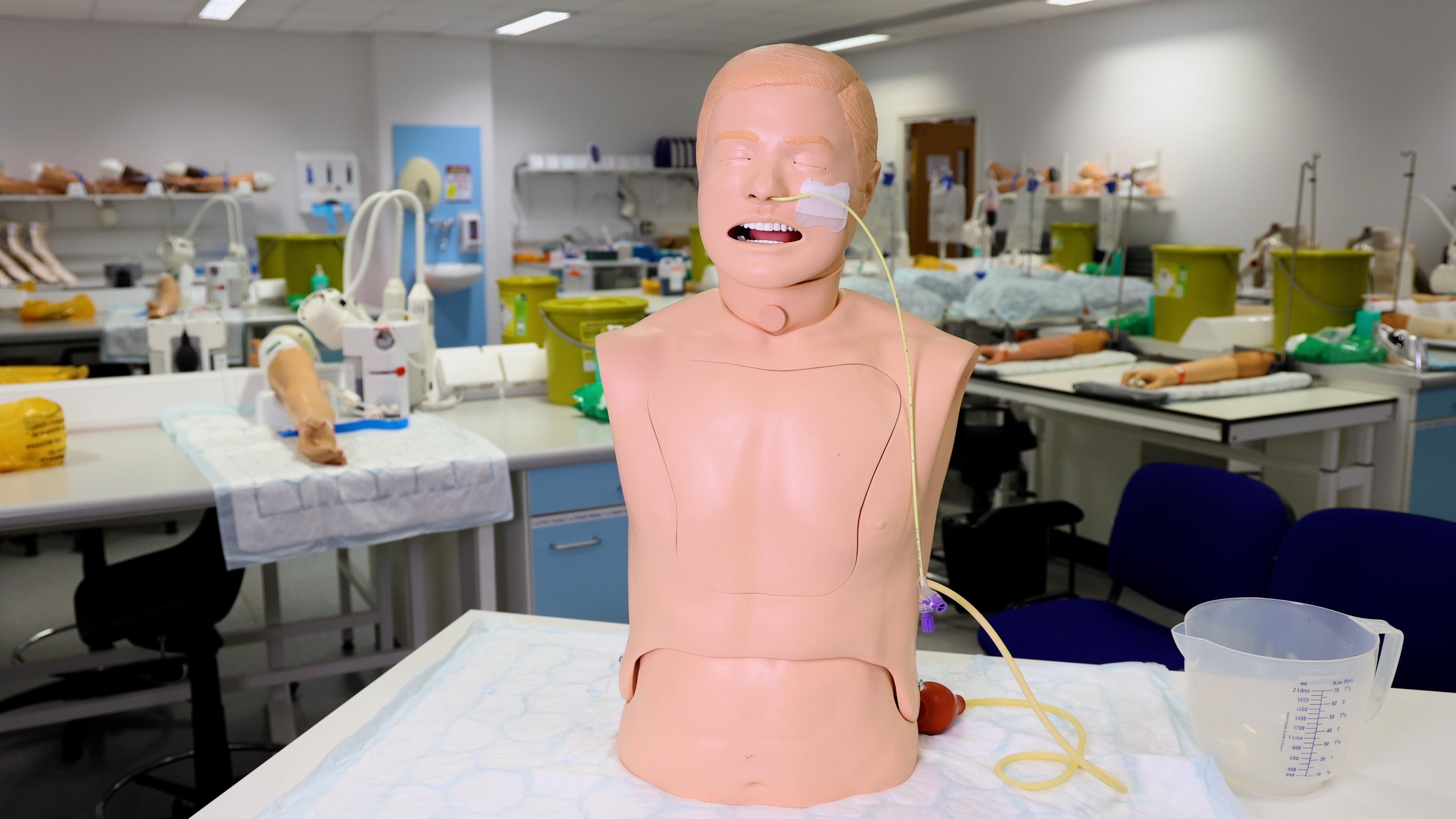 A medical training mannequin with an NG (nasogastric) tube inserted. The mannequin is positioned in a clinical training lab with medical equipment and other training tools in the background.