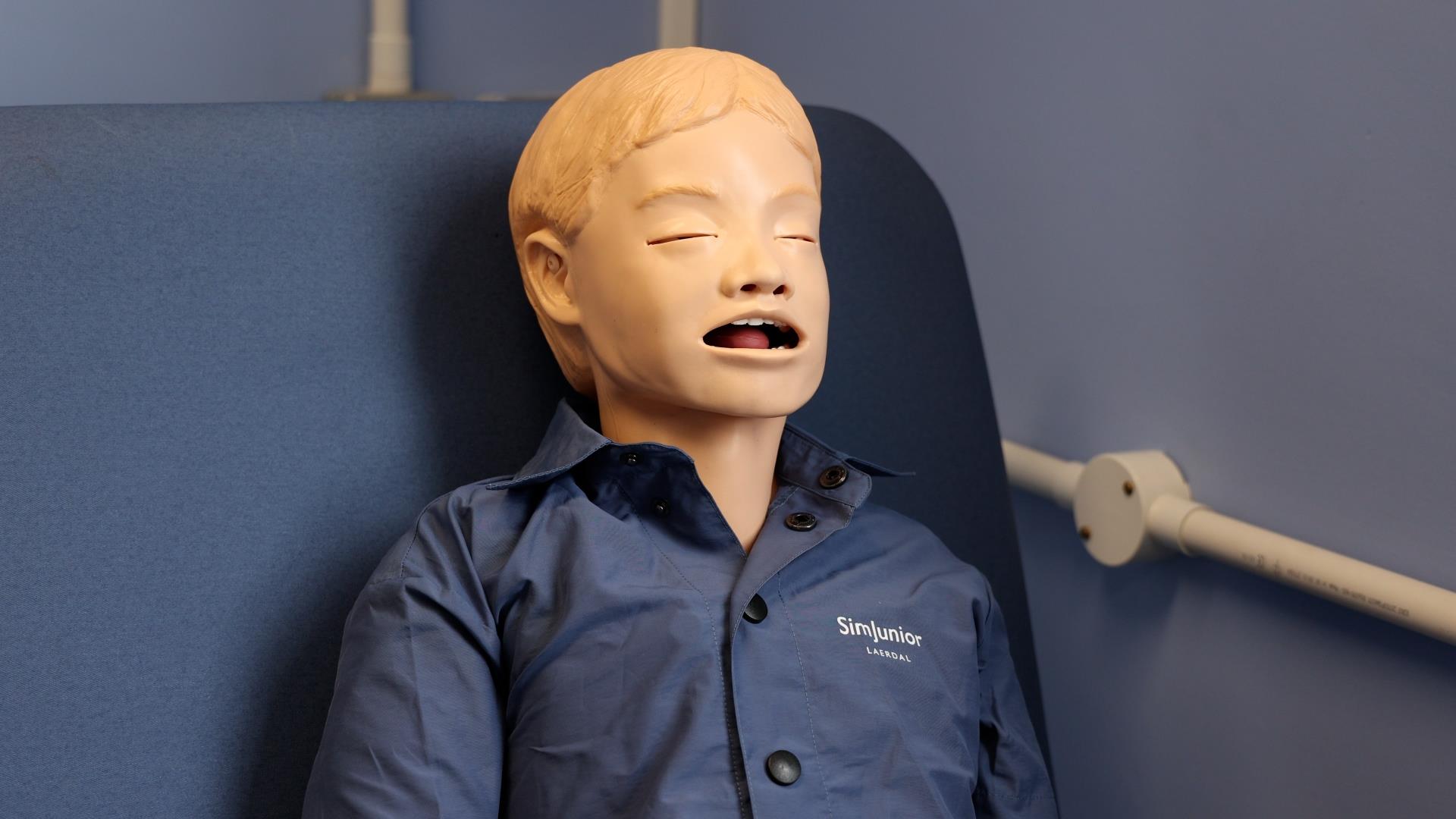 “A pediatric training mannequin, wearing a blue shirt with ‘SimJunior’ branding, seated upright in a clinical setting.”