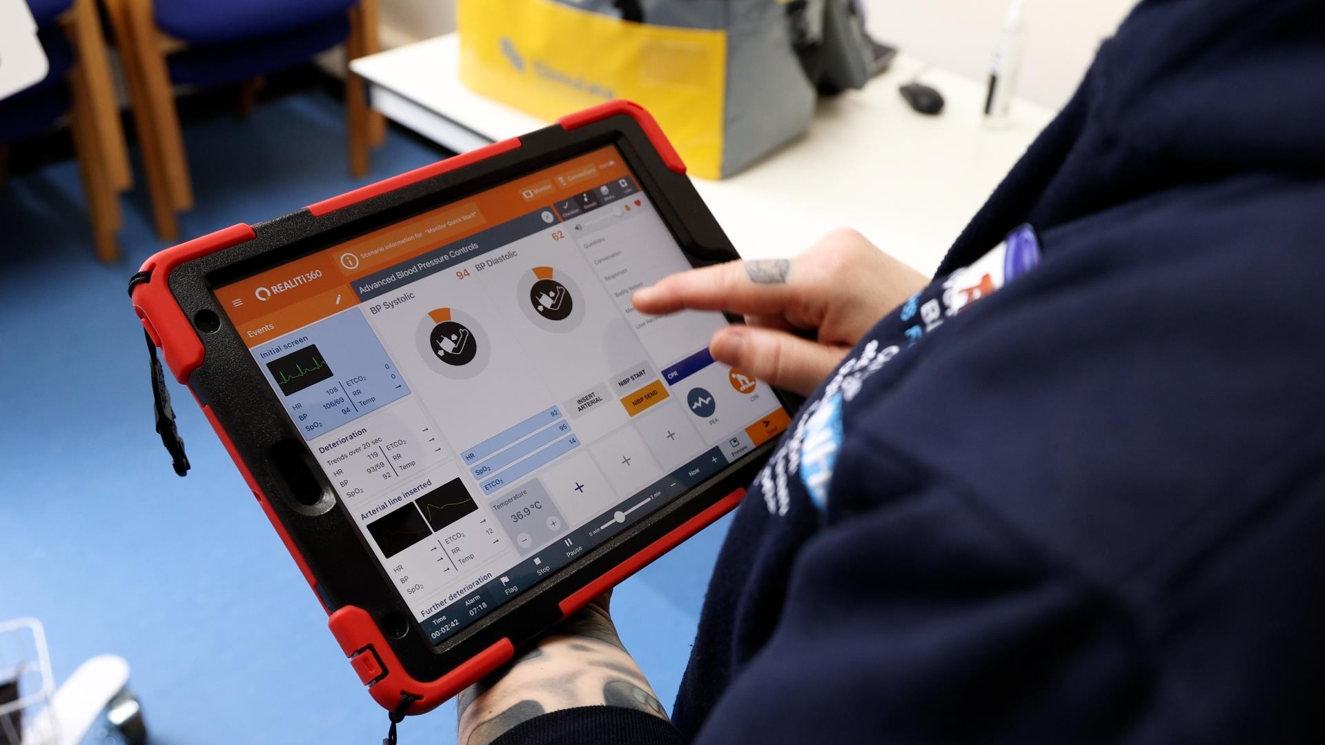 “A person using a tablet device with a medical simulation software interface displayed, showing vital signs and monitoring controls.”